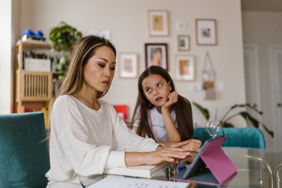 A mother working on her tablet at home while her daughter watches from nearby, illustrating remote work challenges.